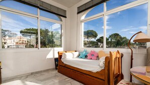 Un dormitorio con cama de madera, una silla, una mesa y ventanas amplias con vistas a una casa y árboles.