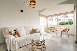 Un salon moderne avec un canapé blanc, une table basse en verre, des fauteuils en osier et une grande fenêtre donnant sur des immeubles.