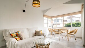 Un salón luminoso con un sofá blanco, una mesa de centro de cristal, sillas de mimbre y un amplio ventanal con vistas a edificios y zonas verdes.