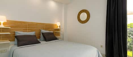 A bedroom with a bed, bedside lamps, a headboard, a wall-mounted mirror, and a window with curtains.