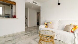 Una sala de estar moderna con un sofá blanco, cojines coloridos, una mesa de centro de mimbre y una zona de cocina con armarios blancos y una cesta roja.