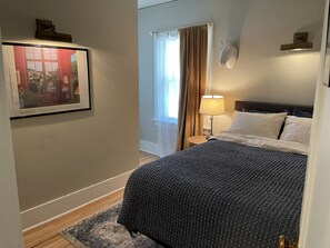 Una habitación de hotel con cama, mesita de noche, lámpara, un cuadro en la pared y ventana con cortinas.