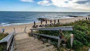En trätrappa leder ner till strandstolar och parasoller, med en sandstrand och havet som bakgrund.