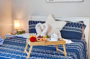 Un lit bien fait, recouvert d’une couvre-lit bleue à motifs, avec un oreiller blanc, un plateau en bois contenant des éléments de petit-déjeuner et une lampe de chevet.