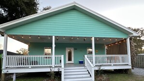 Une maison turquoise avec un porche à l’avant, des balustrades blanches et un petit jardin devant.