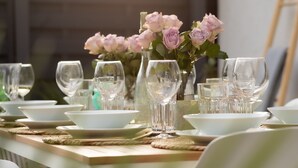 Une table à manger dressée avec de l’assiette, des bols et des verres blancs, agrémentée d’un vase rempli de fleurs roses.