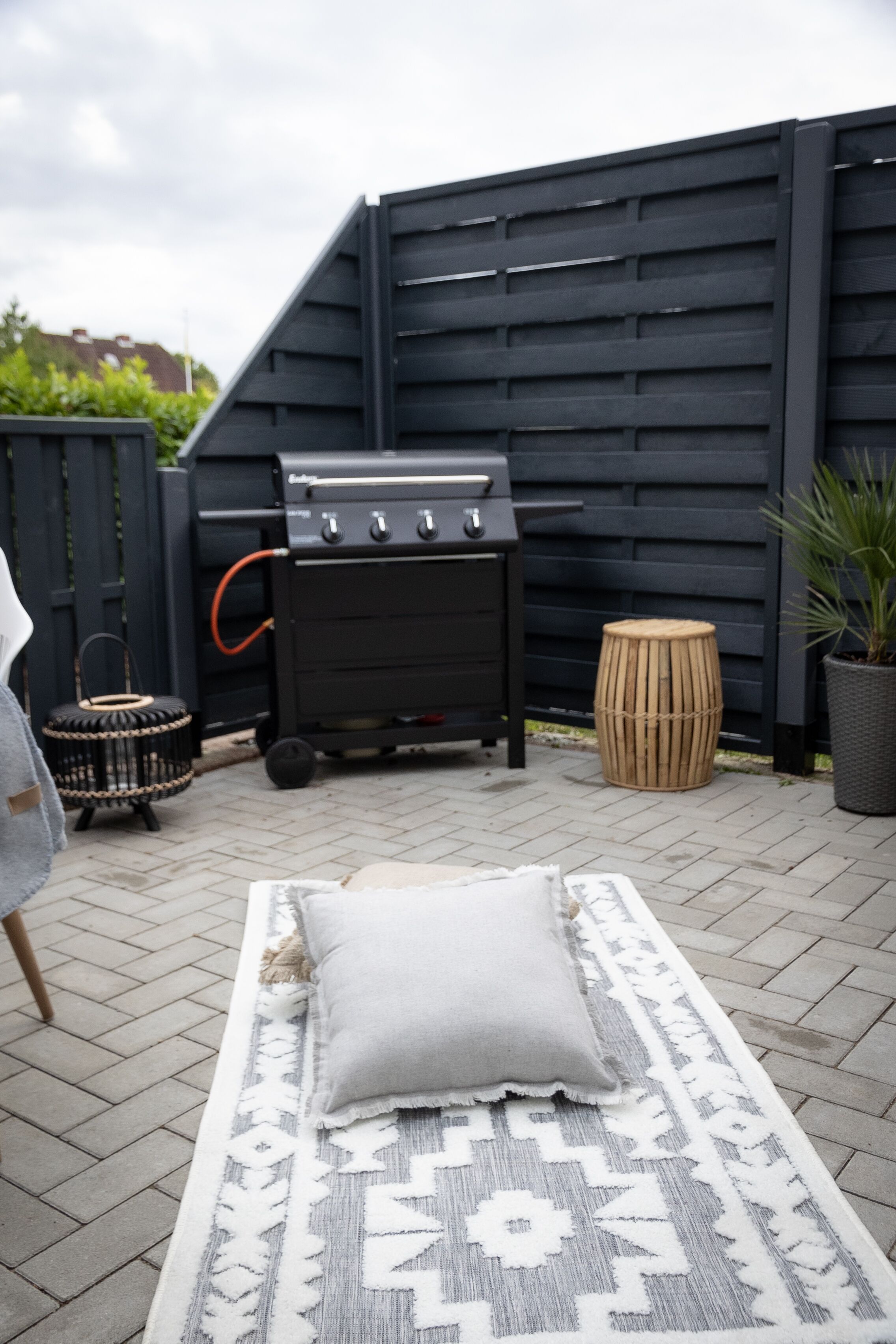 Een terras met een barbecue, een getextureerd vloerkleed, een kussen, een rieten mand en een bloempot.