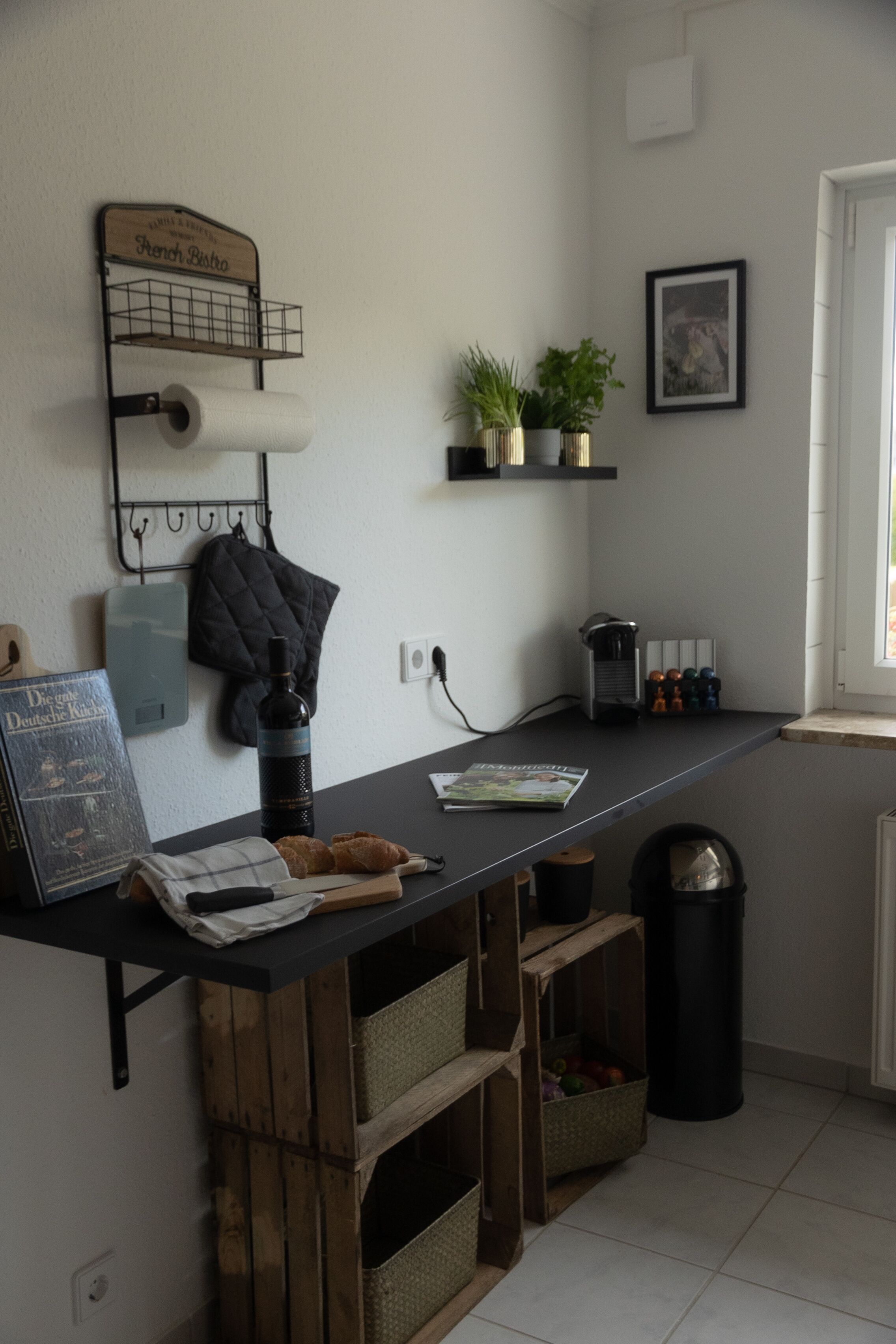 A kitchen with a wooden counter, a hanging rack with kitchen utensils, a potted plant, and a trash bin.