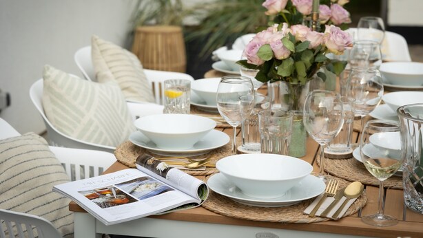 Une table à manger dressée avec de l’assiette blanche, des verres et des couverts, agrémentée d’un magazine et d’un vase rempli de fleurs roses.