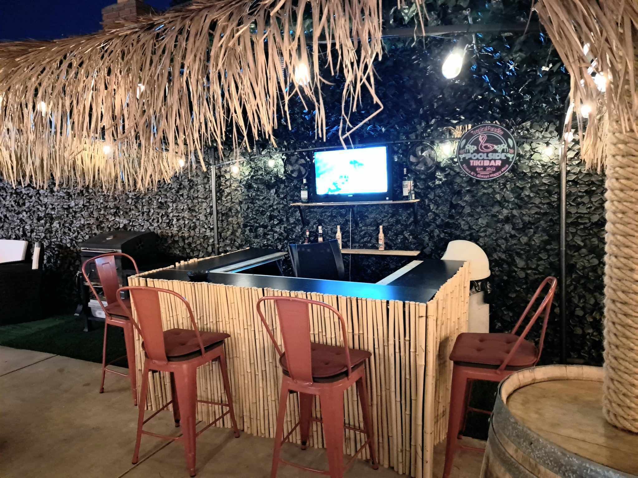 Un espace bar extérieur doté d’un toit de chaume, de chaises en bambou, d’un écran de télévision et d’un mur en pierre servant de décor.