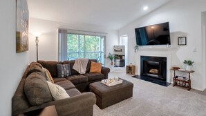 A living room with a large brown sectional sofa, a flat-screen TV mounted above a fireplace, and a large window providing natural light.