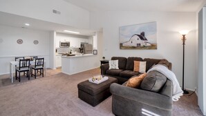 A modern living room with a gray sofa, a dining table with chairs, a kitchen area, and a painting on the wall.