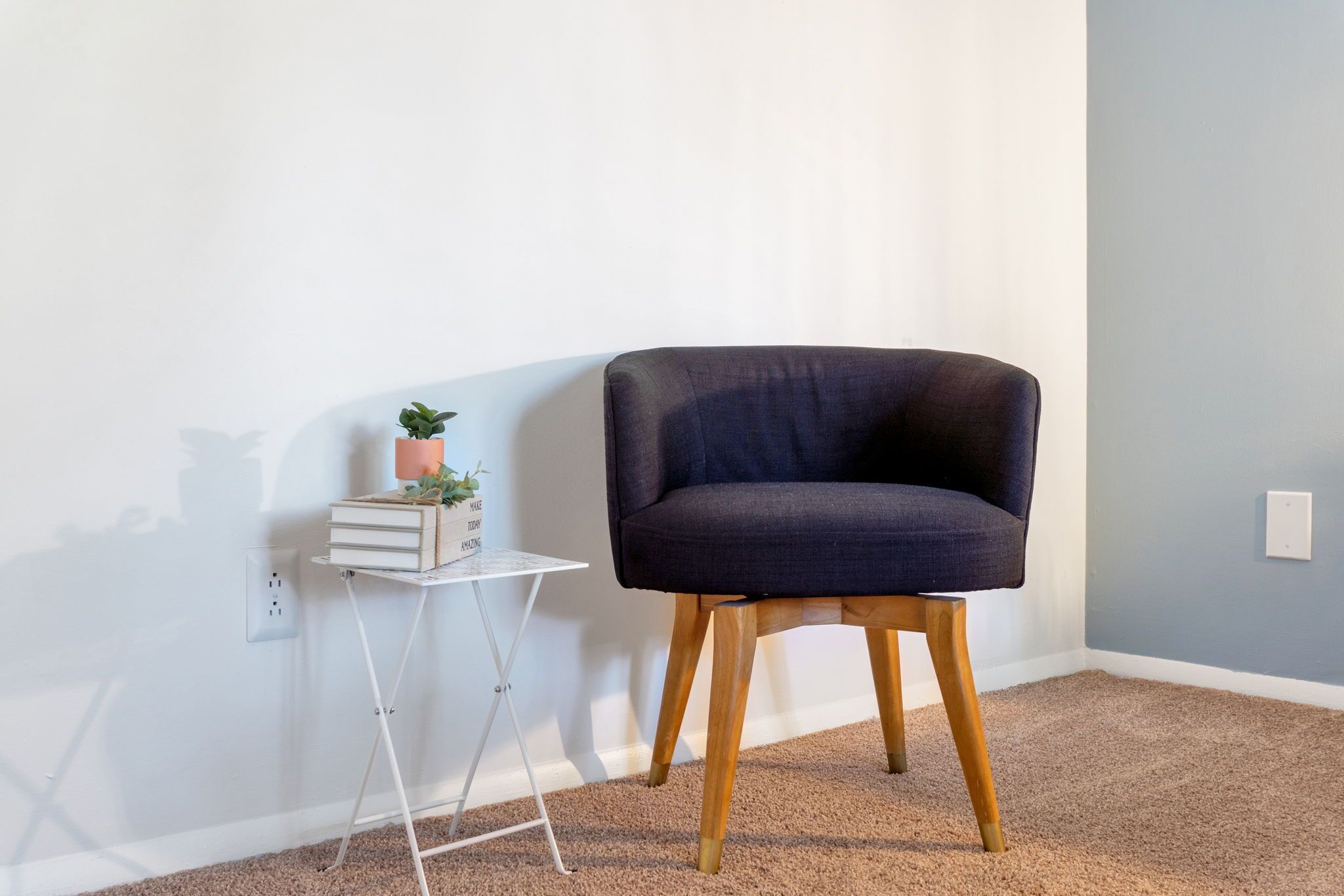 Quarto com uma poltrona estofada azul escura, uma pequena mesa de apoio branca com uma planta, e uma parede branca com tomada.