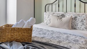 A neatly made bed with floral bedding, a wicker basket with towels, and a gray blanket.