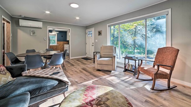 Ein modernes Wohnzimmer mit einer Couch, Sesseln, einem Esstisch und einem großen Fenster mit Blick auf Bäume.