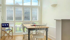 A dining area with a wooden table and white chairs, large windows with blinds, and a view of the outdoors. - 2 bedroom accommodation in Beadnell, nr Chathill (Beadnell, nr Chathill)