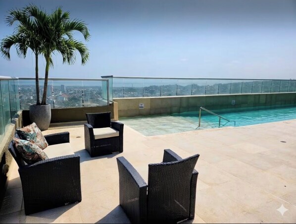 A rooftop pool area with lounge chairs, a palm tree, and a city view.