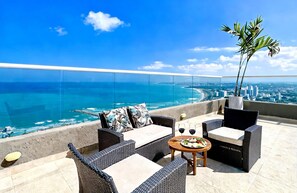 A balcony with wicker furniture, a table with a plate of food and glasses, and a view of the ocean and city skyline.
