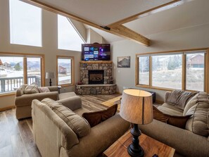 A cozy living room with a stone fireplace, large windows, and a flat-screen TV mounted above it. - Overlook, gorgeous large home with Hot Tub and Pool Table, 5/3, near Granby Ranch - Sleeps 12 (Granby)