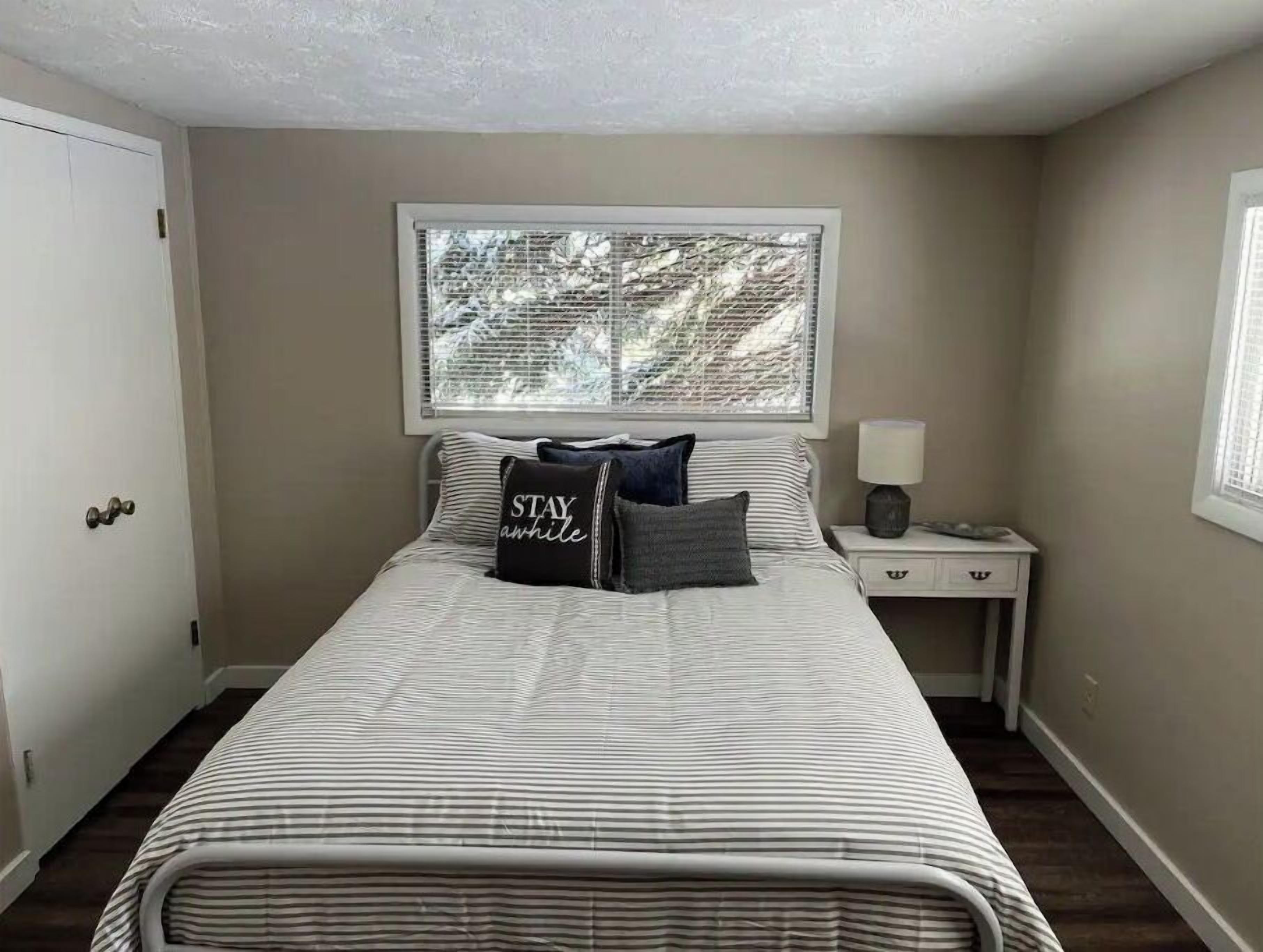 Um quarto com cama, janela com persianas, mesa de cabeceira e abajur, e uma porta.