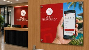 A hotel reception area with a wooden reception desk, a red wall with the hotel's logo, and a poster of a smartphone displaying a booking interface. - Casa Família: Praia, Campeche e Conforto (Florianópolis)