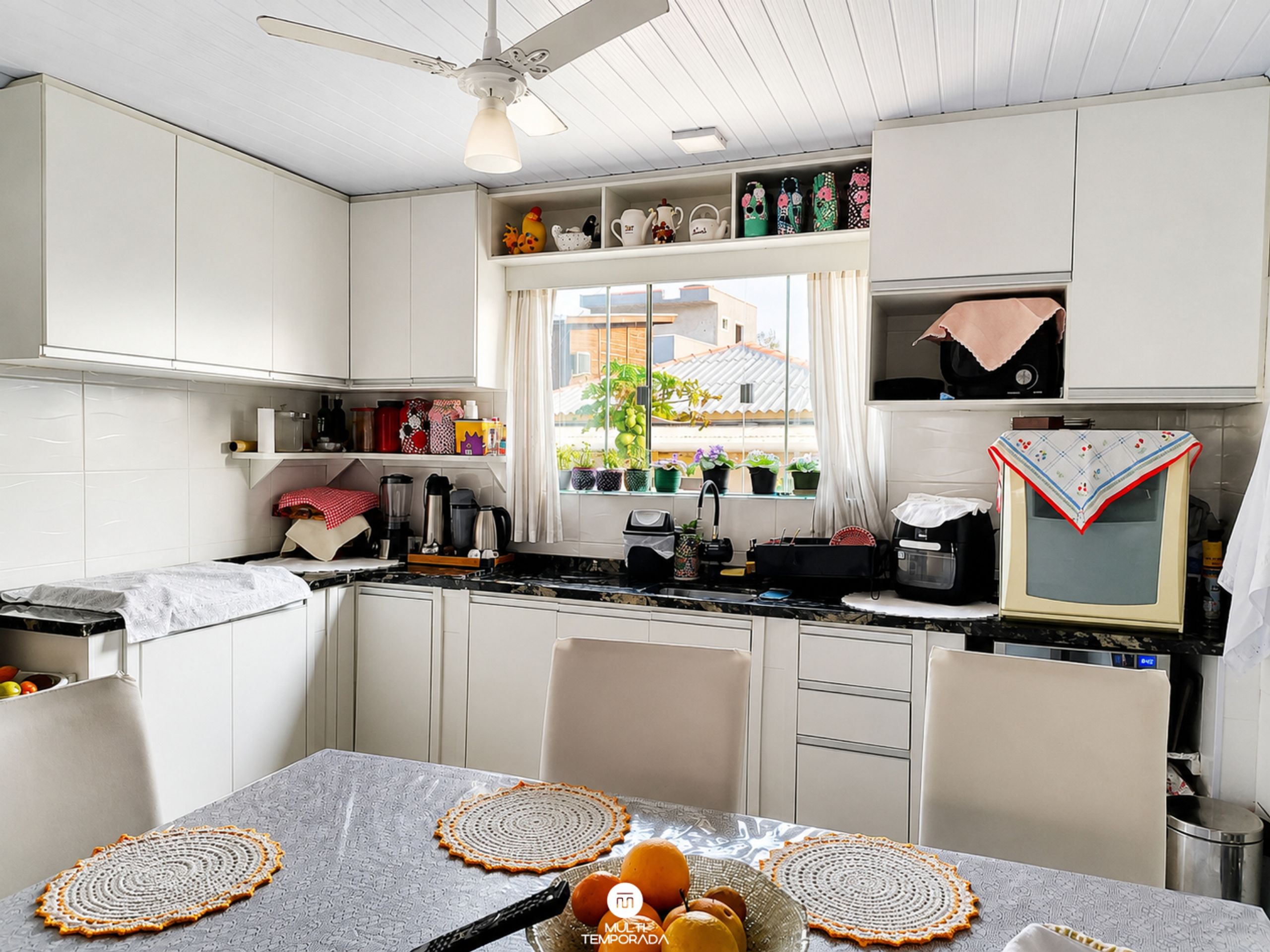 Cocina compacta con armarios blancos, una ventana con plantas en macetas y una zona de comedor con una mesa para dos.
