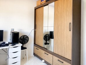 A room with a wooden wardrobe, a desk with a computer and two fans, and a chair.