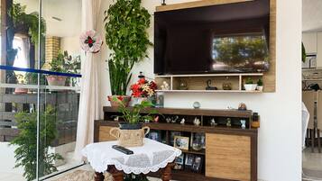 A modern living room with a flat-screen TV mounted on the wall, a wooden entertainment unit below, and a glass partition with plants.