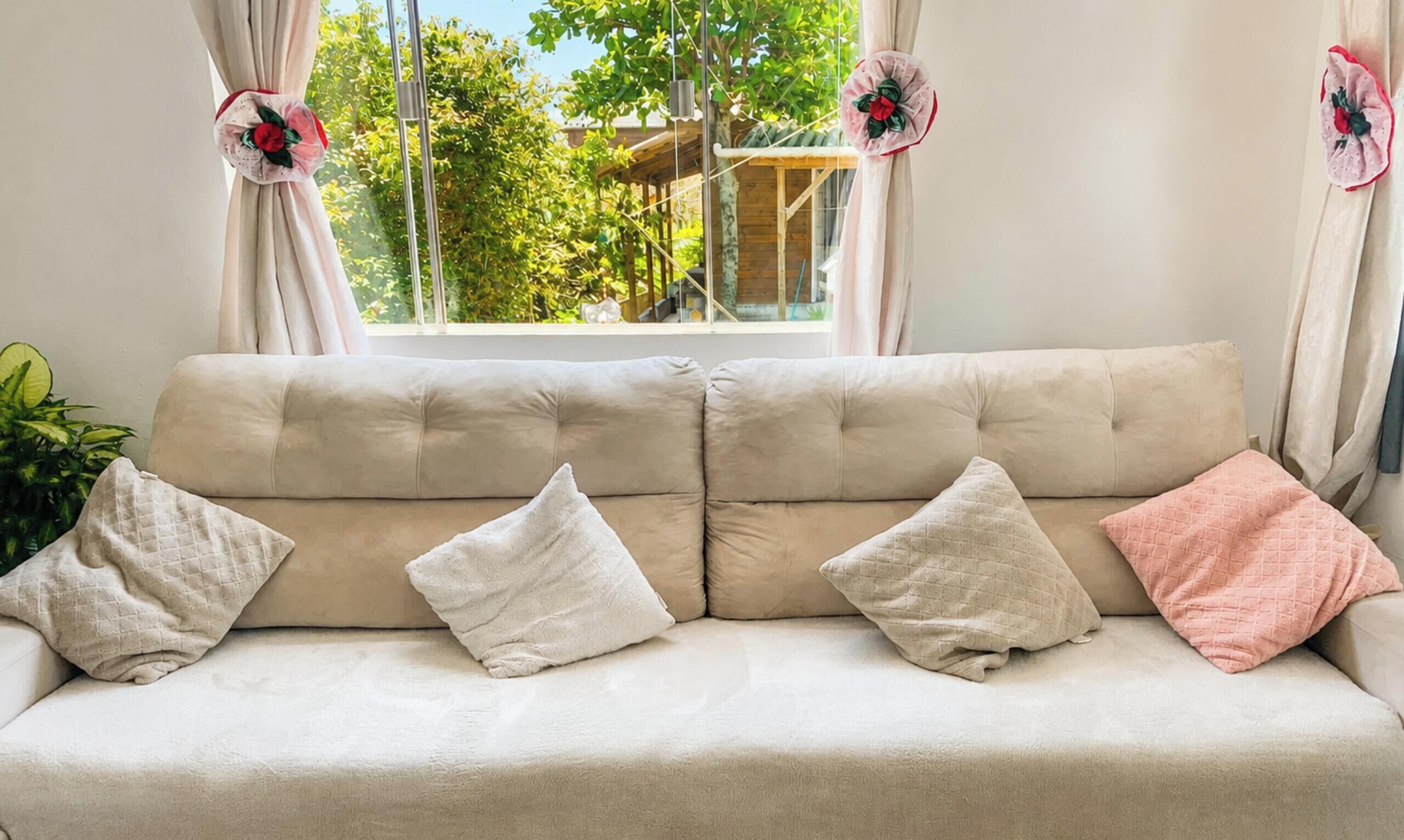 A beige sofa with four cushions, a window with curtains, and a view of greenery outside.