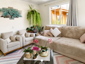 Una sala de estar con un sofá color beige, dos sillones, una mesa de centro con plantas y un ventanal con cortinas.