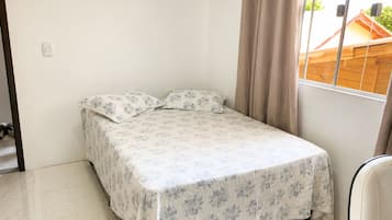 Quarto pequeno, com uma cama de solteiro, colcha florida, cabeceira de madeira e janela com cortinas.