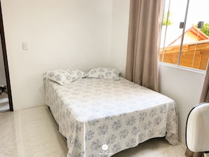 A small, single-bed room with a floral bedspread, a wooden headboard, and a window with curtains.