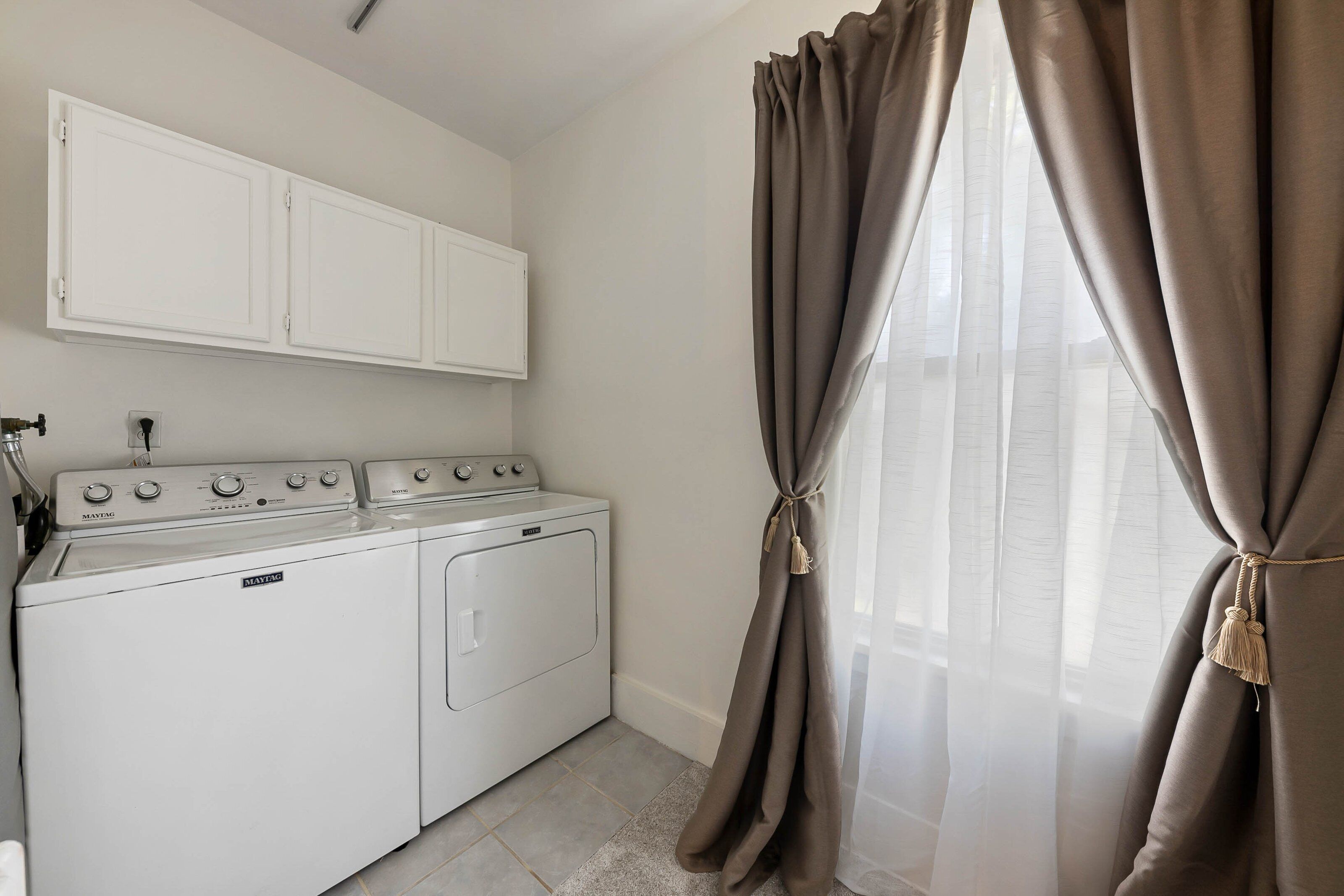 A laundry room with a washing machine and dryer, a white cabinet above, and a window with beige curtains.
