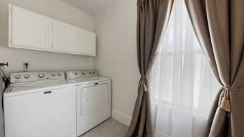 A laundry room with a washing machine and dryer, a white cabinet above, and a window with beige curtains.