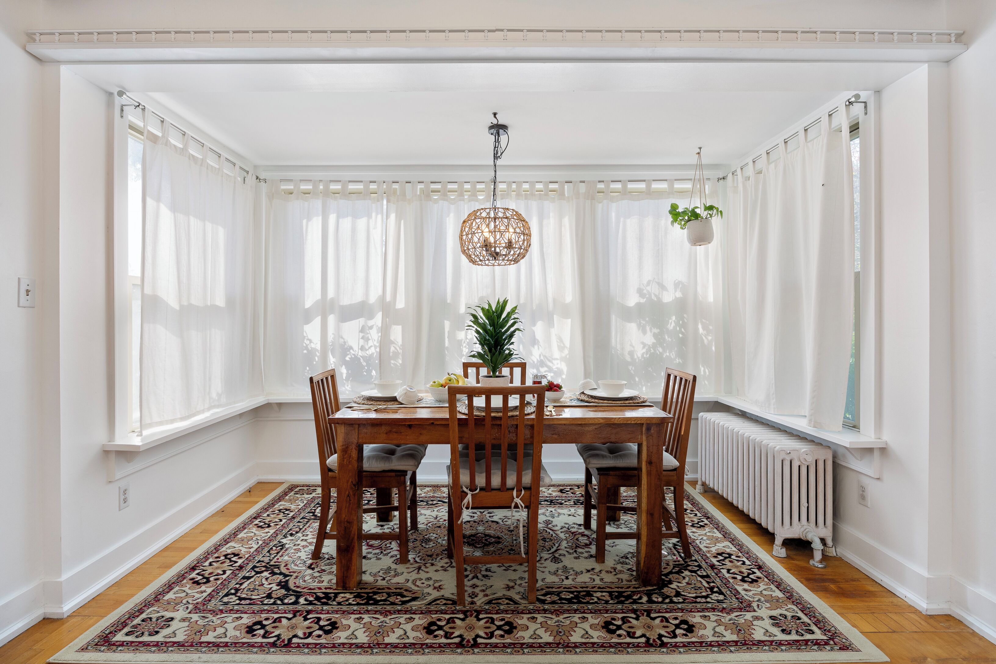 Une salle à manger avec une table en bois, quatre chaises, un tapis à motifs, un luminaire suspendu et une plante en pot.