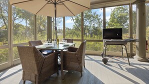 Een overdekt terras met rieten stoelen, een tafel met glazen en een fles, en een barbecue.
