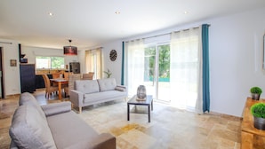 A spacious living area with a sofa, dining table, and chairs, a kitchen in the background, and large windows with curtains. - Cozy home in Grignan with private swimming pool, can be inside or outside (Grignan)