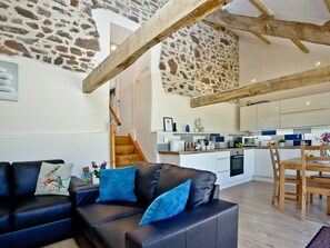 Un espace de vie moderne avec un mur en pierre, des poutres en bois, un canapé avec des coussins bleus, un coin repas et une cuisine dotée de meubles blancs.