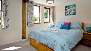 A bedroom with a wooden bed frame, a floral bedspread, a painting on the wall, and a window with curtains.