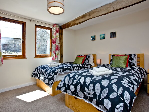 A bedroom with two beds, a window, a wooden beam ceiling, and a view of a stone building outside. - 2 bedroom accommodation in Umberleigh (Umberleigh)