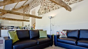 A modern living room with a stone wall, wooden beams, a black leather sofa, and a wooden floor. - 2 bedroom accommodation in Umberleigh (Umberleigh)