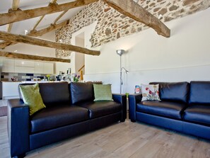 A modern living room with a stone wall, wooden beams, a black leather sofa, and a wooden floor. - 2 bedroom accommodation in Umberleigh (Umberleigh)