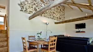 A dining area with a stone wall, wooden ceiling beams, a TV mounted on the wall, and a black sofa in the living area. - 2 bedroom accommodation in Umberleigh (Umberleigh)