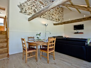 A dining area with a stone wall, wooden ceiling beams, a TV mounted on the wall, and a black sofa in the living area. - 2 bedroom accommodation in Umberleigh (Umberleigh)