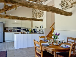 A dining area with a wooden table set for two, a kitchen in the background, and exposed wooden beams. - 2 bedroom accommodation in Umberleigh (Umberleigh)