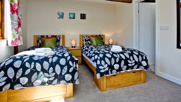 A room with two wooden beds, each with a leaf-patterned bedspread, a wooden ceiling beam, and a small wall-mounted light fixture. - 2 bedroom accommodation in Umberleigh (Umberleigh)