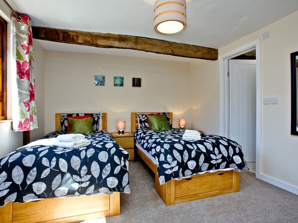 Quarto com duas camas de madeira, cada uma com colcha estampada em folha, uma viga de madeira no teto e uma luminária pequena fixada na parede.