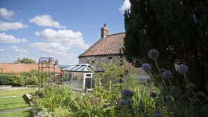 Een stenen huis met een pannendak, een glazen conservatorium en een tuin vol planten en bloemen.