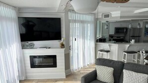 Un salon moderne avec un téléviseur à écran plat, une cheminée, un canapé gris et un coin cuisine à l’arrière-plan.