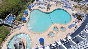 Une grande piscine extérieure divisée en plusieurs zones, entourée de chaises longues et de parasols, et située à proximité d’une plage.